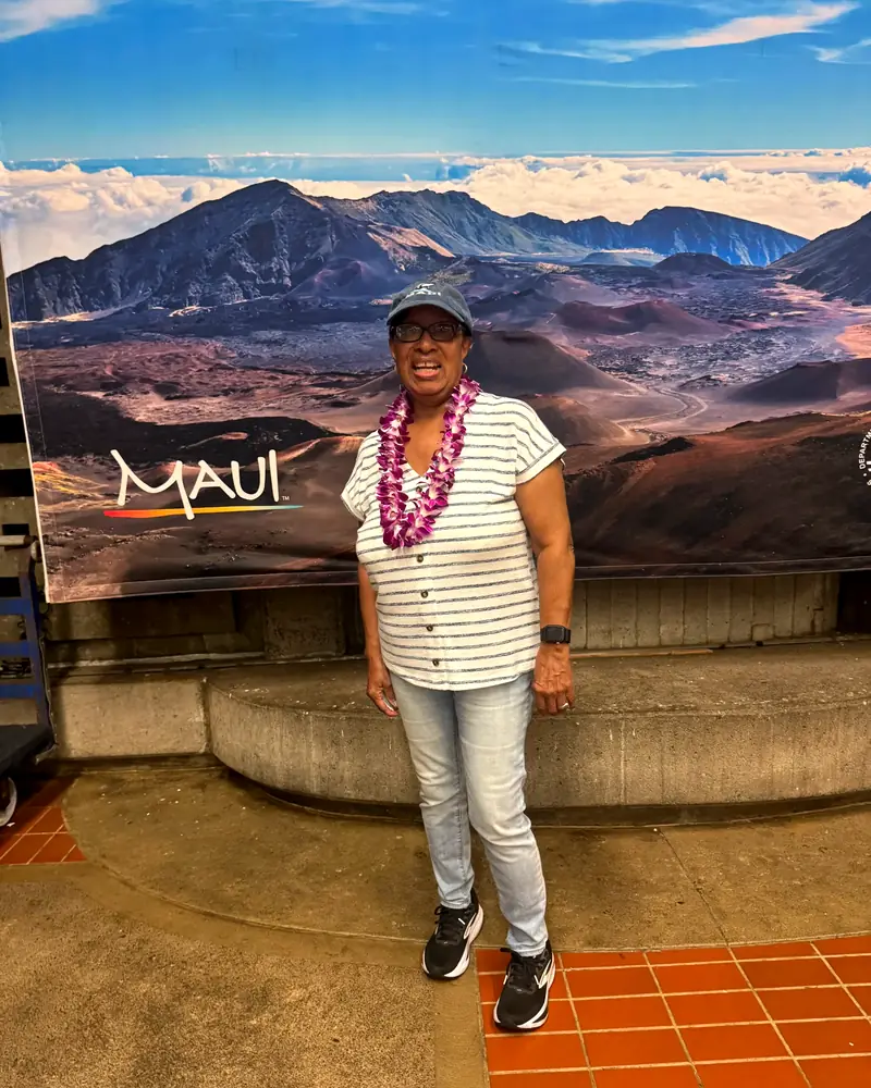 A smiling traveler wearing a fresh orchid lei in front of the Maui mural