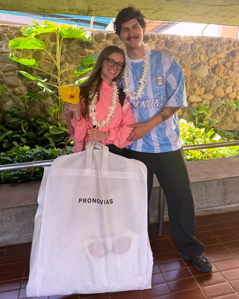Couple wearing orchid lei on arrival, carrying a wedding dress garment bag