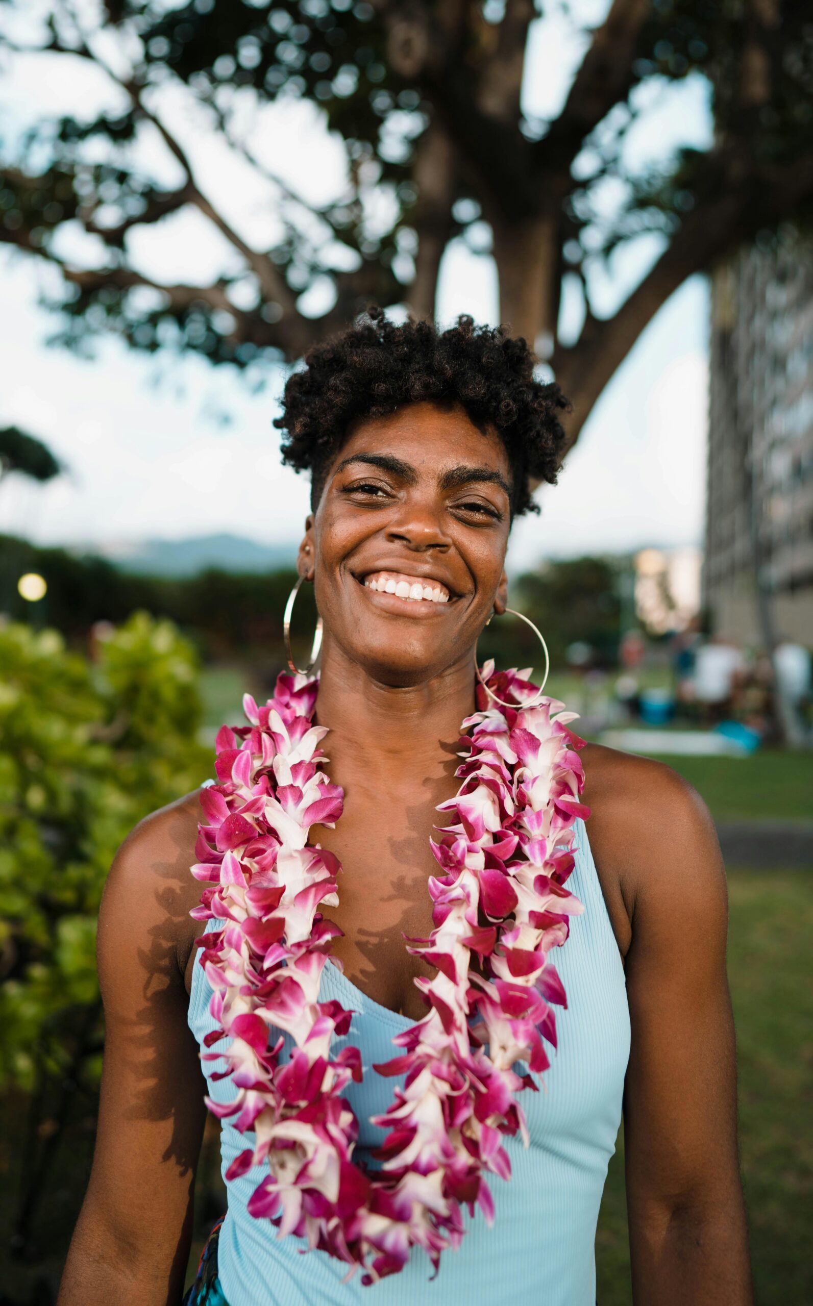 Cheerful African American female in traditional leis flower garland and big earrings in local green yard with bushes and trees